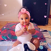 Djéliya a rejoint le concours — aidez-le/la à gagner de superbes lots ! baby, infant, child, toy, blanket, headband, pink, heart_effects, floor, indoor, smiling, cute, sitting, colorful, playful, happy, person, clothing, appliance, floor_tiles