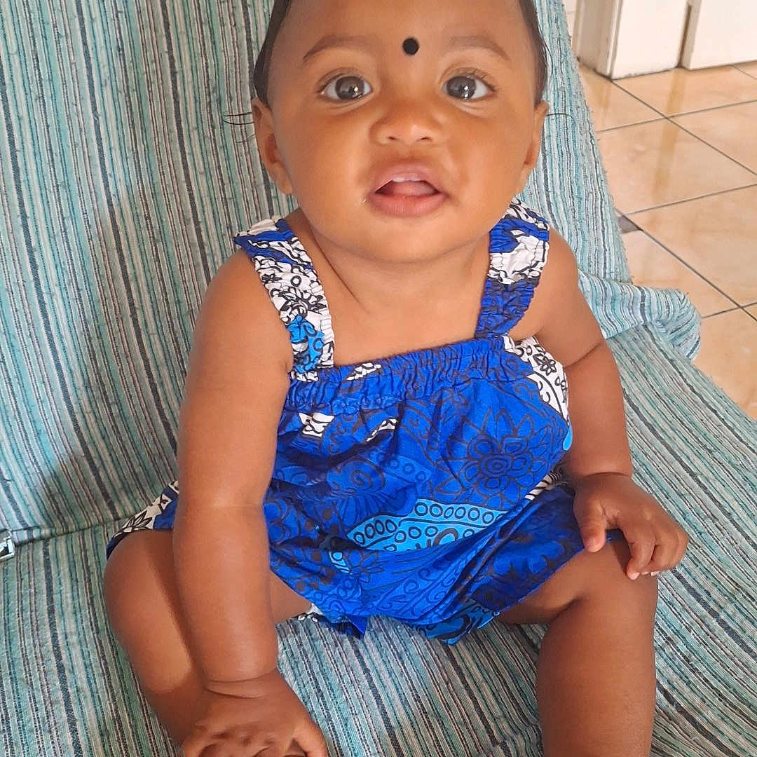 Djéliya a rejoint le concours — aidez-le/la à gagner de superbes lots ! baby, black_dot, blue_dress, child, curious, eyes, face, floor, floor_tiles, footwear, hand, indoor, indoors, patterned_clothing, person, sitting, striped_chair, toddler, white_sandals, young_child