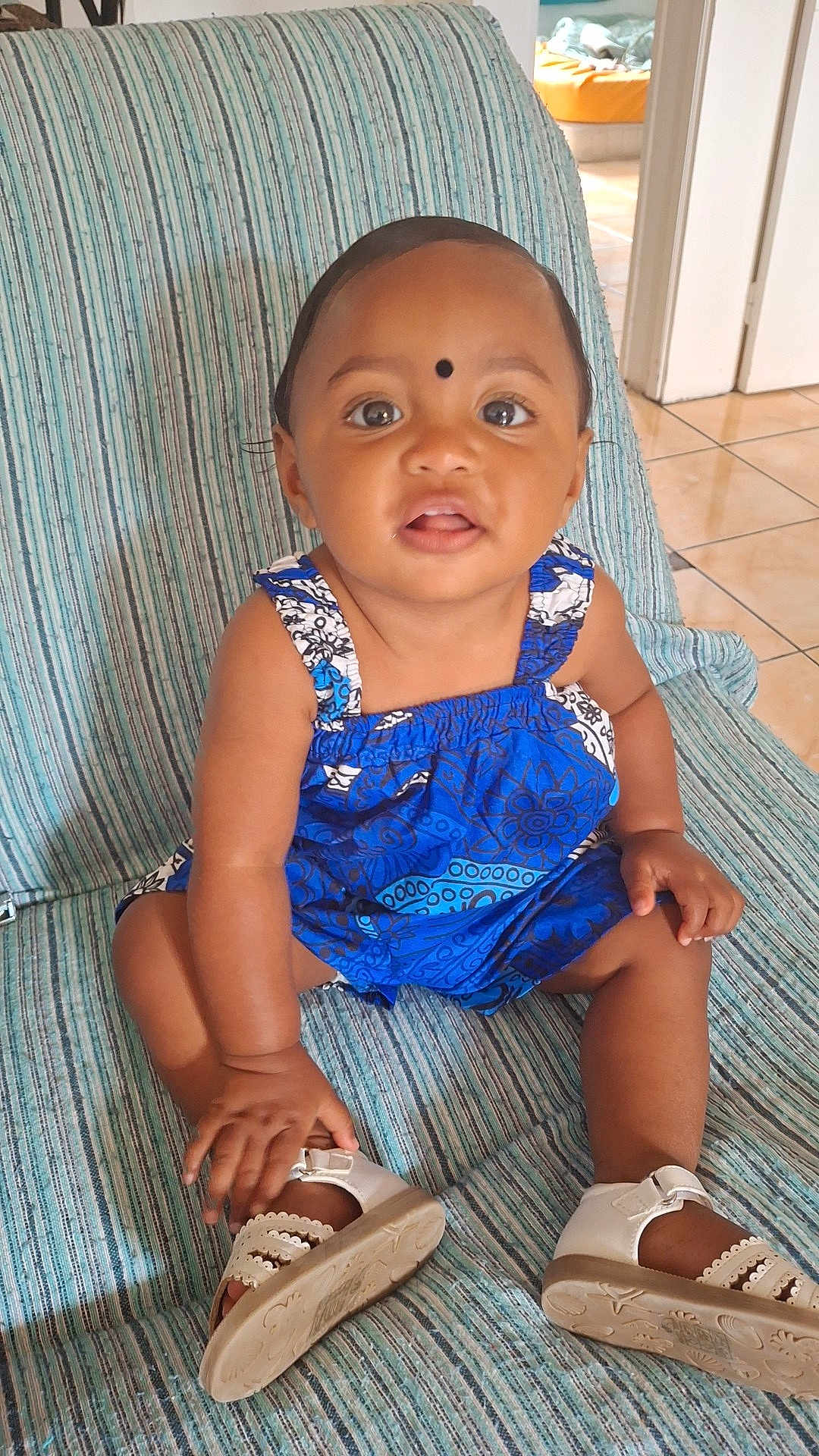 Djéliya a rejoint le concours — aidez-le/la à gagner de superbes lots ! toddler, child, blue_dress, striped_chair, white_sandals, indoor, floor_tiles, curious, sitting, hand, face, eyes, black_dot, patterned_clothing, baby, footwear, person, young_child, indoors, floor