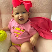 Athalia is registered to the contest to win money with this photo: baby, bunny_slippers, cape, child, cute, footwear, happy, headband, indoors, infant, onesie, pink_bow, playful, portrait, seated, smiling, soft_chair, superhero_costume, toy, yellow_ball