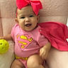baby, bunny_slippers, cape, child, cute, footwear, happy, headband, indoors, infant, onesie, pink_bow, playful, portrait, seated, smiling, soft_chair, superhero_costume, toy, yellow_ball