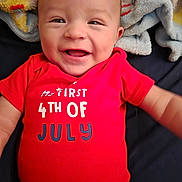 Liam joined the competition — help win amazing prizes! american, baby, blanket, celebration, child, close_up, clothing, cute, face, happy, holiday, indoor, infant, lying_down, person, portrait, red_onesie, smiling, soft_texture, text_on_clothing