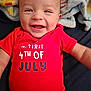 baby, smiling, red_onesie, text_on_clothing, blanket, infant, face, happy, portrait, lying_down, close_up, child, cute, clothing, soft_texture, indoor, celebration, holiday, american, person