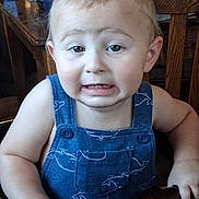 Milo is registered to the contest to win money with this photo: toddler, child, baby, portrait, close_up, face, sitting, high_chair, blue_overalls, clothing, blond_hair, eyes, expression, frowning, indoor, dining_room, table, chair, wooden_furniture, photograph