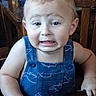 toddler, child, baby, portrait, close_up, face, sitting, high_chair, blue_overalls, clothing, blond_hair, eyes, expression, frowning, indoor, dining_room, table, chair, wooden_furniture, photograph