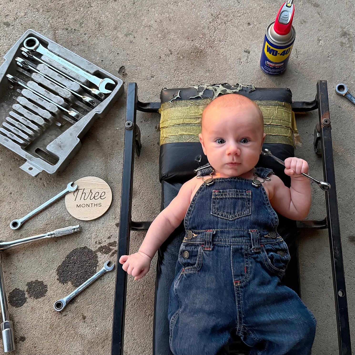 Gage is registered to the contest to win money with this photo: baby, denim_overalls, wrench, tools, wd40, mechanic_creeper, concrete_floor, oil_spots, wooden_sign, three_months, infant, workshop, mechanic, face, person, laying_down, small_tool, metal_tools, floor, cute
