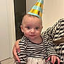 baby, celebration, child, closeup, curious_expression, cute, door, dress, face, floor_tiles, hand, holding, indoor, long_hair, party_hat, patterned_clothing, person, striped_clothing, striped_shirt, white_wall