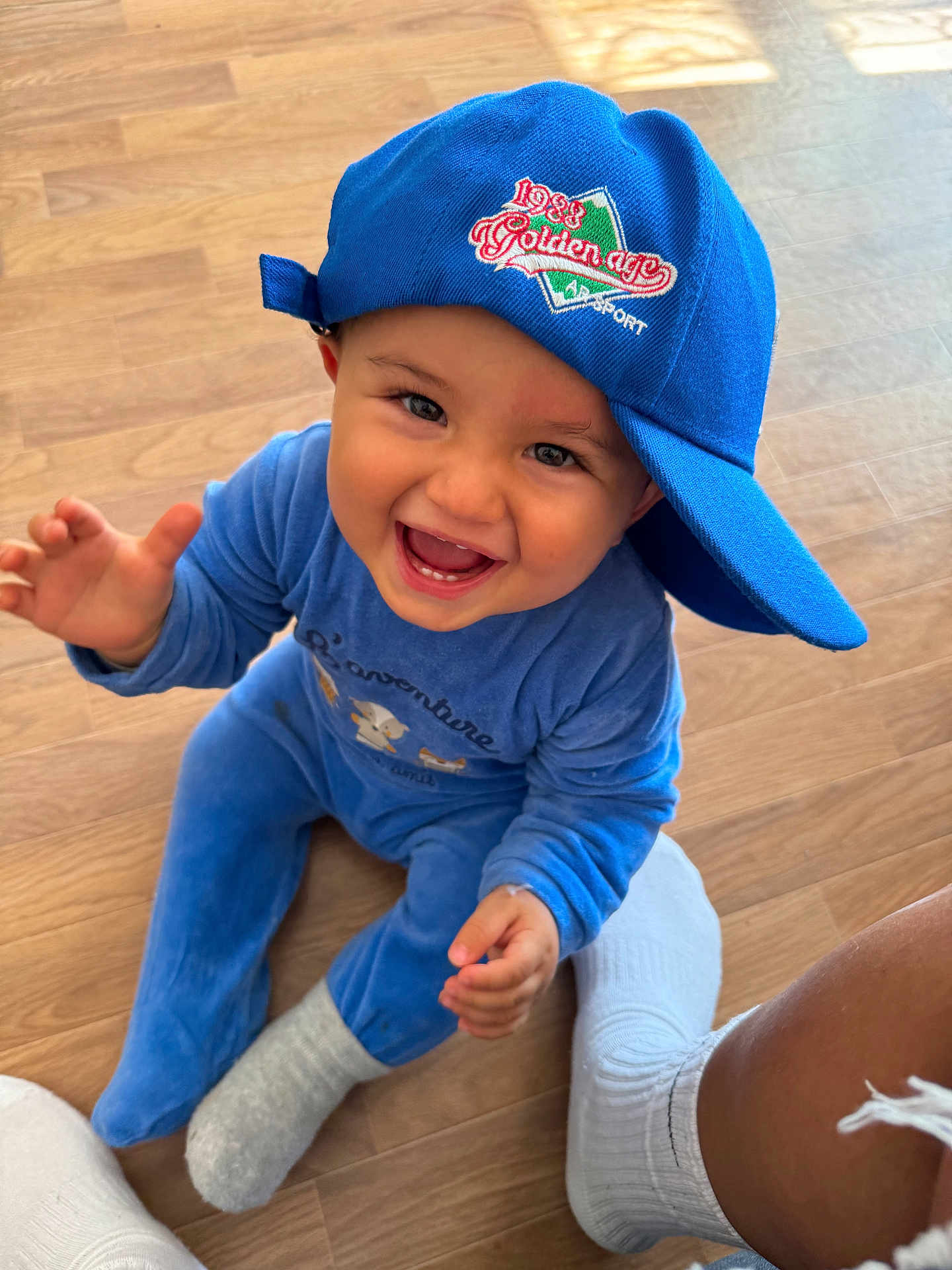 Menzo participe au concours pour gagner de l'argent avec cette photo : baby, child, blue_cap, blue_onesie, smiling, happy, sitting, wooden_floor, indoor, sock, ripped_jeans, person, cute, playful, portrait, casual, infant, reaching_out, hat, joyful