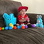 baby, child, red_cap, red_shirt, brown_couch, easter_eggs, blue_bunny_bucket, yellow_chick_toy, blue_bunny_plush, plastic_eggs, indoor, floor, wooden_baseboard, sitting, cute, toy, colorful, curious, person, play