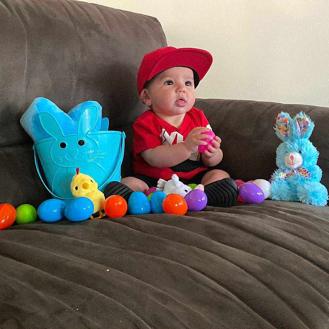 Donald Scudder Jr is registered to the contest to win money with this photo: baby, blue_bunny_bucket, blue_bunny_plush, brown_couch, child, colorful, curious, cute, easter_eggs, floor, indoor, person, plastic_eggs, play, red_cap, red_shirt, sitting, toy, wooden_baseboard, yellow_chick_toy