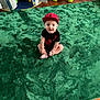 baby, child, red_cap, black_clothing, green_carpet, wooden_floor, play_tent, toy_snake, toy_storage, floor, indoor, smiling, sitting, cute, happy, portrait, person, play_area, kid, headwear
