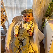 Ismael participe au concours pour gagner de l'argent avec cette photo : baby, child, smiling, hat, bow_tie, suspenders, shirt, pants, barefoot, indoor, chair, blanket, plant, wood_floor, window, curtain, wicker_basket, happy, portrait, cozy
