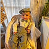 baby, child, smiling, hat, bow_tie, suspenders, shirt, pants, barefoot, indoor, chair, blanket, plant, wood_floor, window, curtain, wicker_basket, happy, portrait, cozy