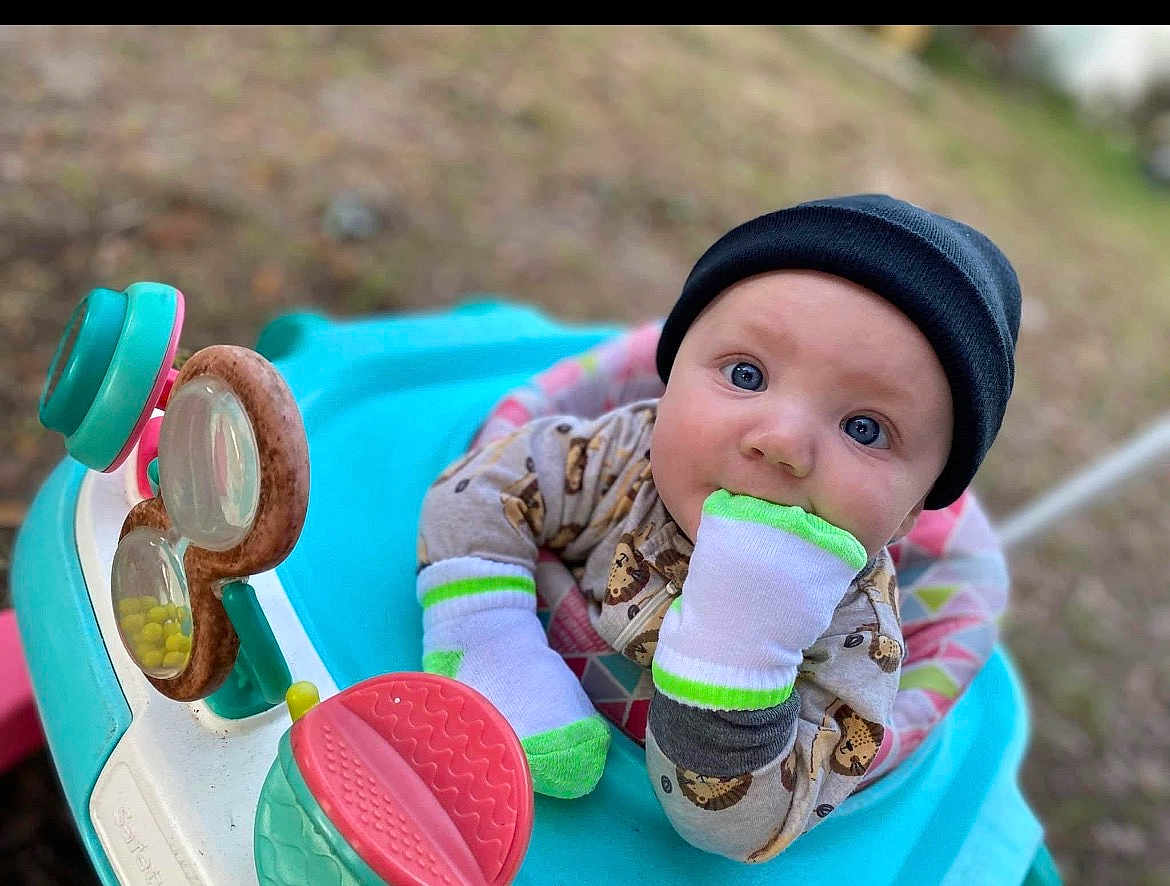 Christian is registered to the contest to win money with this photo: baby, infant, child, face, blue_eyes, beanie, mittens, sock, teether, toy, activity_seat, walker, outdoor, bokeh_background, grass, portrait, closeup, gaze, playful, patterned_pajamas