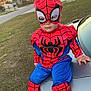 child, toddler, costume, spiderman_costume, mask, face, sneakers, car_trunk, car, grass, lawn, suburban, driveway, sunset, portrait, sitting, outdoor, holiday_costume, street, playful