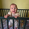 Genevieve is registered to the contest to win money with this photo: baby, infant, child, smiling, standing, crib, nursery, portrait, indoor, home, hands, face, eyes, open_mouth, clothing, onesie, barefoot, bedding, furniture, happy