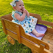 Clara is registered to the contest to win money with this photo: baby, child, outdoor, wagon, wooden_wagon, grass, sunlight, dress, bow, smile, happy, infant, greenery, summer, playful, cute, sitting, portrait, nature, childhood
