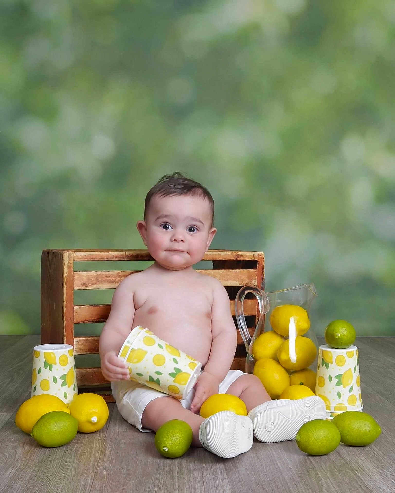 Karam is registered to the contest to win money with this photo: baby, child, lemon, lime, cup, pitcher, wooden_floor, crate, fruit, sitting, white_shoes, white_shorts, curious_expression, indoor, still_life, portrait, colorful, fresh, playful, smiling