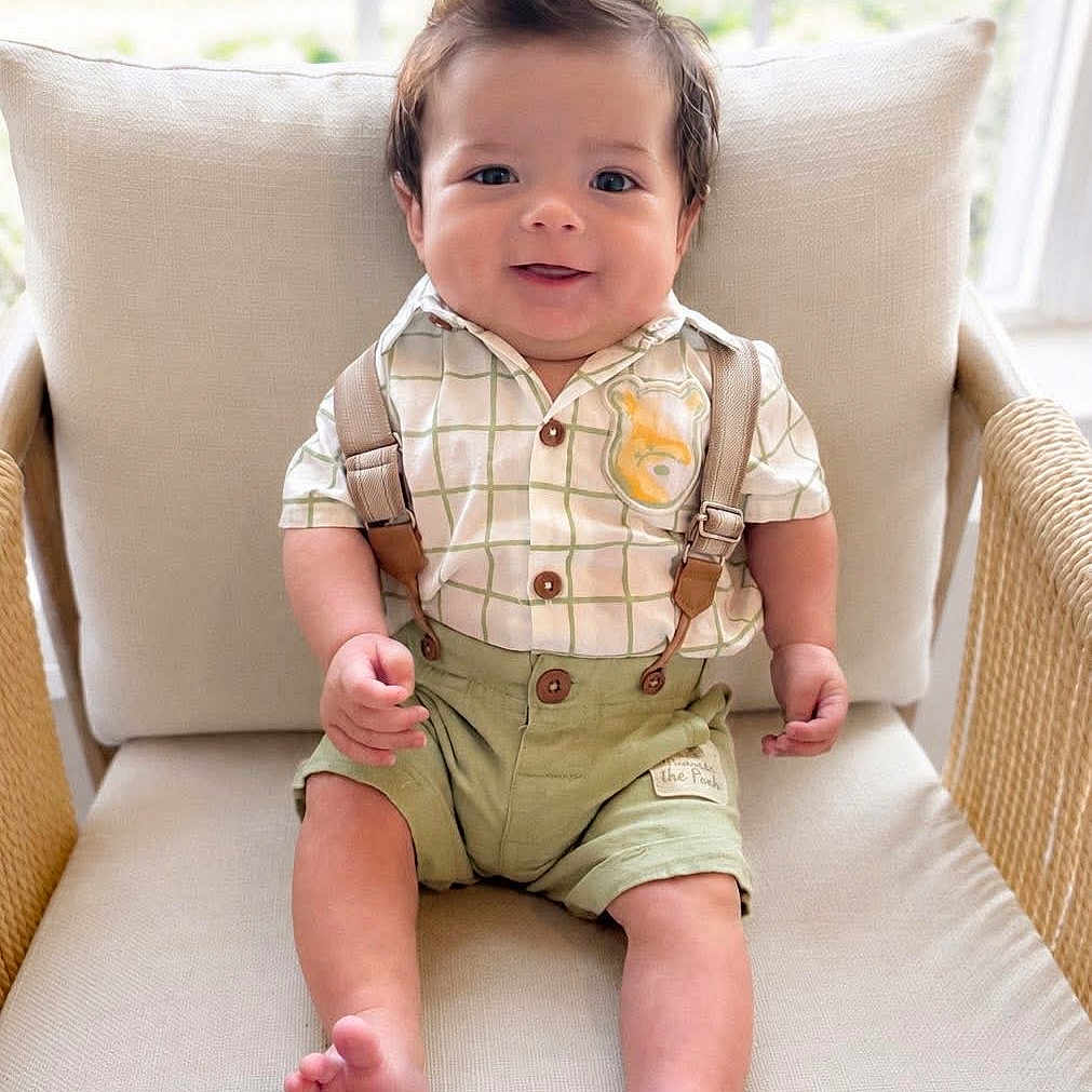Ezaia is registered to the contest to win money with this photo: baby, child, smiling, sitting, indoors, chair, cushion, clothing, suspenders, shorts, hairstyle, window, natural_light, cute, portrait, feet, happy, person, infant, home