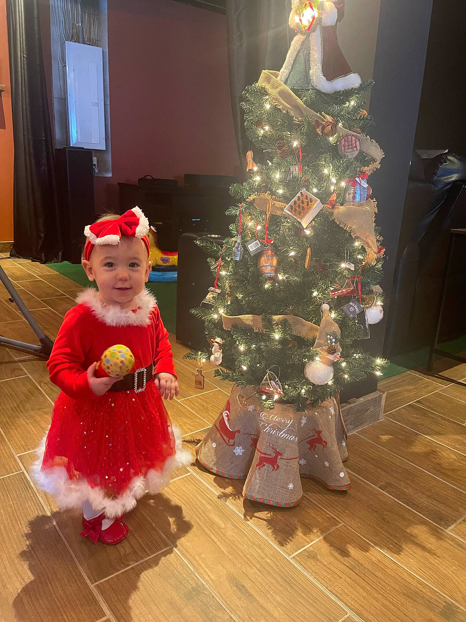 Ellianna is registered to the contest to win money with this photo: baby_toddler_clothing, christmas, christmas_decoration, christmas_eve, christmas_ornament, christmas_tree, door, event, flooring, fun, hardwood, headwear, holiday, holiday_ornament, ornament, person, plant, standing, toddler, tree