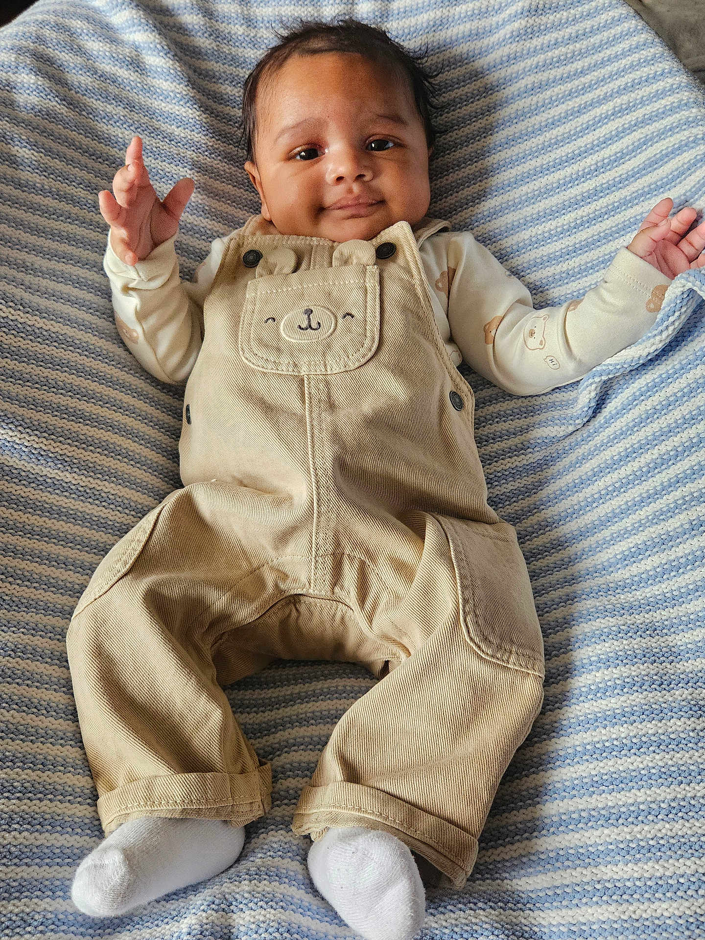 Kairi is registered to the contest to win money with this photo: baby, child, infant, overalls, beige_clothing, white_socks, striped_blanket, smiling, happy, cute, lying_down, small_hands, face, portrait, soft_texture, cozy, indoors, casual, adorable, young