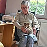 man, person, puppy, dog, pet, sitting, chair, glasses, shirt, jeans, office, home, window, sunlight, printer, desk, cabinet, radiator, indoors, portrait