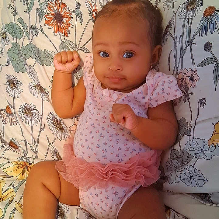Alayah is registered to the contest to win money with this photo: baby, bedspread, child, cute, expression, face, floral_pattern, hair_bow, happy, indoors, infant, lying_down, onesie, pink_clothing, portrait, raised_fists, ruffles, skin, small_hands, socks