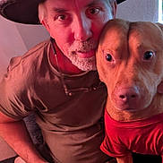 Sonny Boy is registered to the contest to win money with this photo: animal, beard, camouflage_pants, casual_clothing, closeup, companion, dog, face, friendship, hat, human, indoor, looking_at_camera, man, paw, pet, portrait, red_shirt, seated, warm_lighting