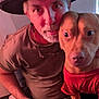 animal, beard, camouflage_pants, casual_clothing, closeup, companion, dog, face, friendship, hat, human, indoor, looking_at_camera, man, paw, pet, portrait, red_shirt, seated, warm_lighting