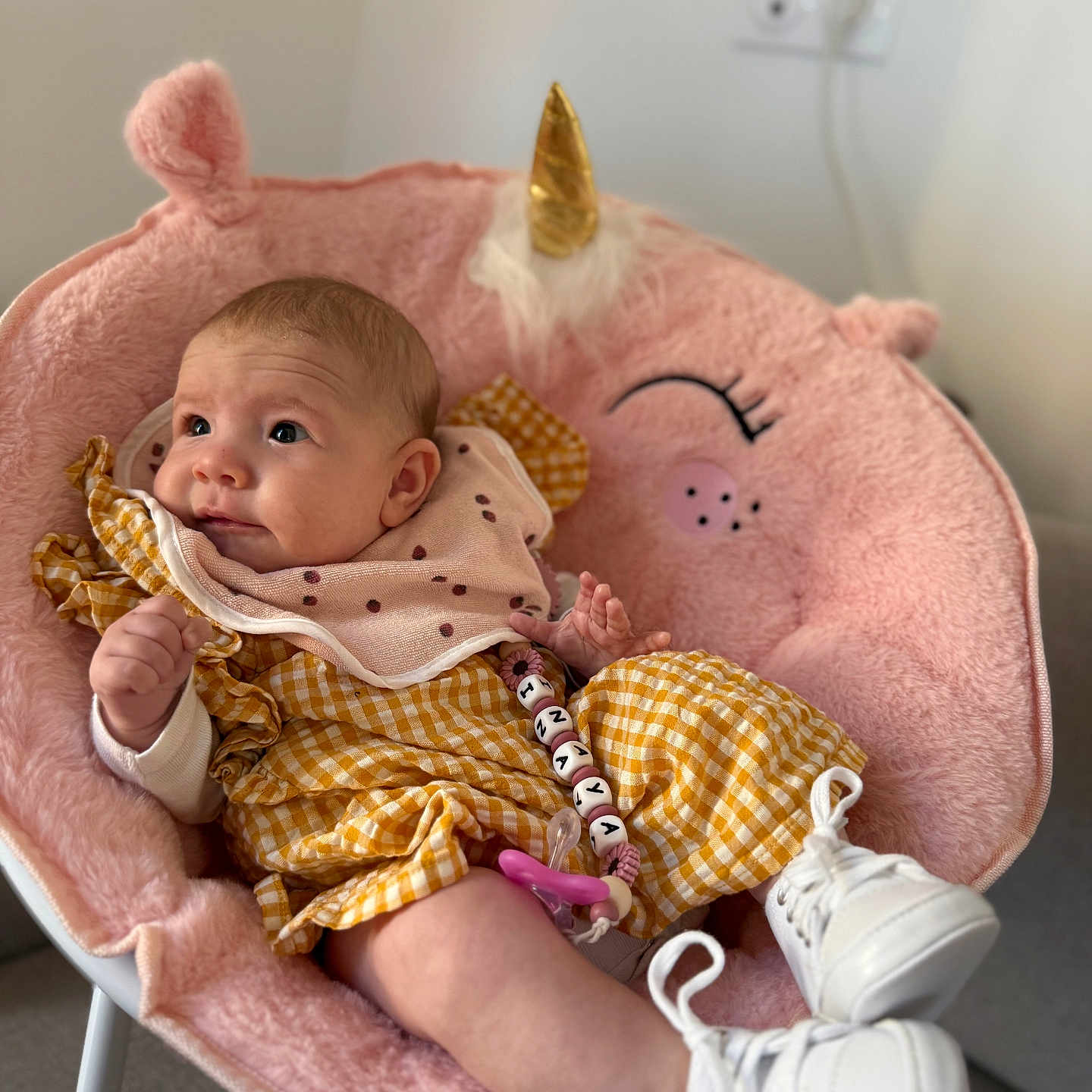 Inaya participe au concours pour gagner de l'argent avec cette photo : baby, baby_accessories, baby_fashion, child, curious_expression, cute, human, indoor, infant, pacifier, pacifier_clip, pink_bib, plush_chair, portrait, room, seated, soft_texture, unicorn_chair, white_shoes, yellow_checkered_clothing
