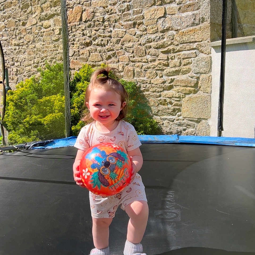 Leïla a rejoint le concours — aidez-le/la à gagner de superbes lots ! baby, ball, child, daylight, footwear, greenery, hair_tie, happy, outdoor, person, play, shadow, shirt, shorts, smiling, socks, stone_wall, sunlight, toddler, trampoline