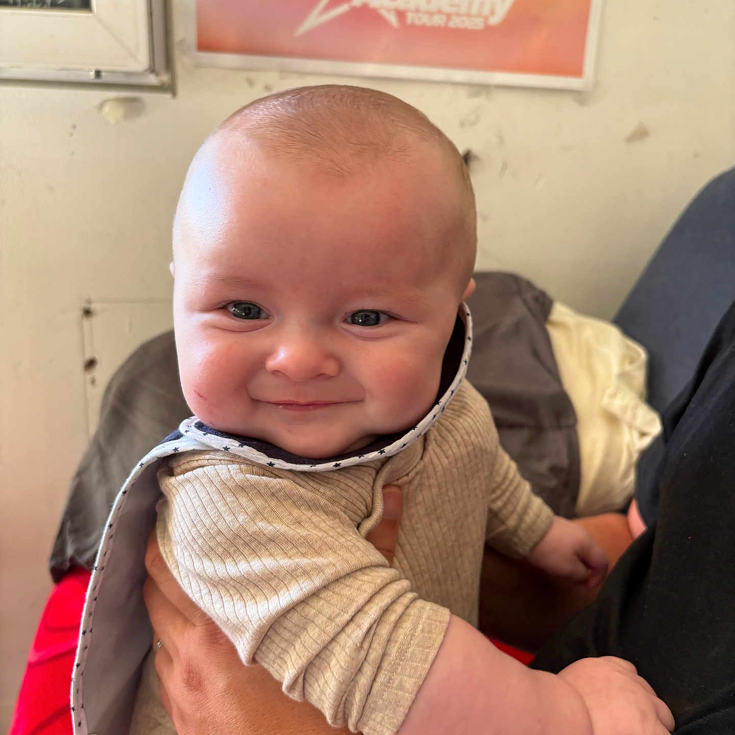 Maël a rejoint le concours — aidez-le/la à gagner de superbes lots ! adult_hand, baby, bib, cheeks, clothing, cute, expression, face, hand, happy, holding, indoor, infant, person, portrait, poster, sitting, skin, smile, wall