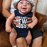 Romeo is registered to the contest to win money with this photo: baby, smiling, hat, onesie, text_on_clothing, adult, tattoo, arm, hand, chair, leather, indoor, person, casual_clothing, happy, infant, sitting, footwear, sock, parent
