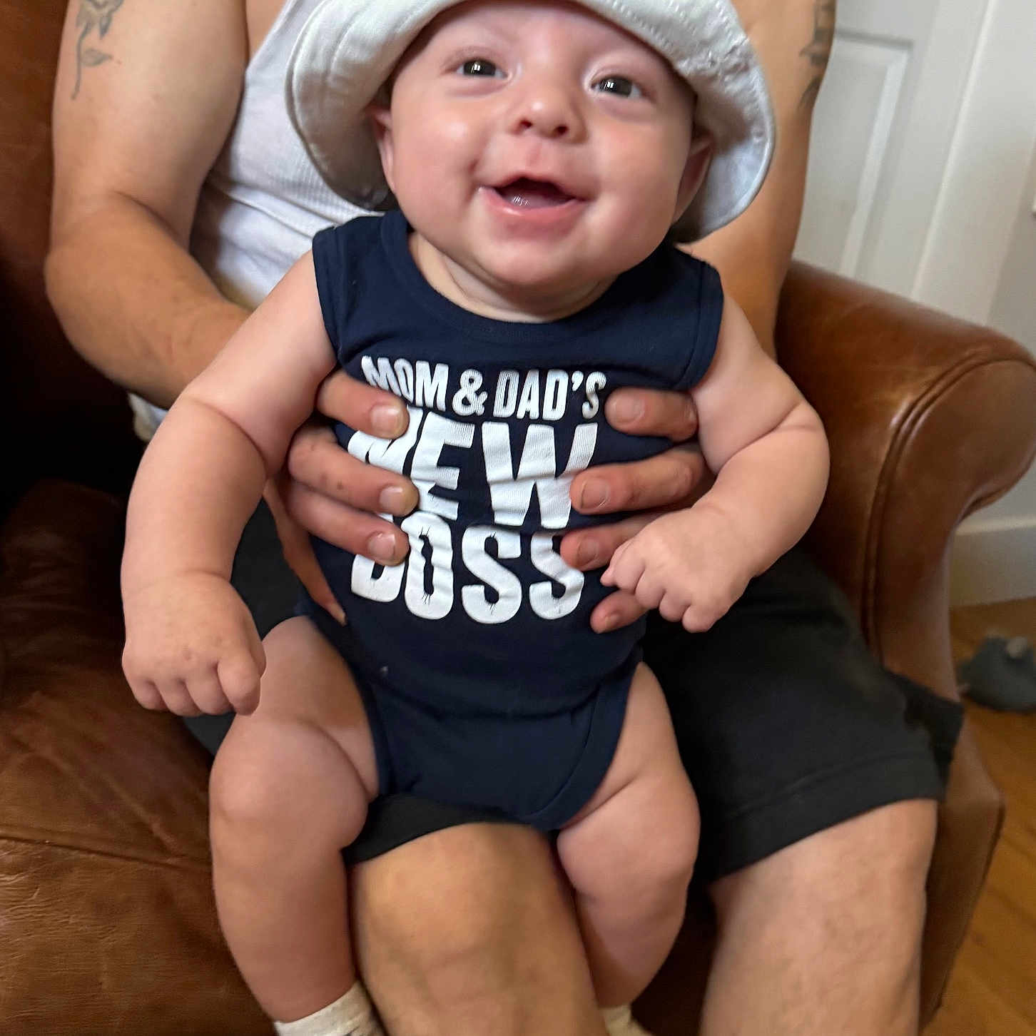 Romeo is registered to the contest to win money with this photo: baby, smiling, hat, onesie, text_on_clothing, adult, tattoo, arm, hand, chair, leather, indoor, person, casual_clothing, happy, infant, sitting, footwear, sock, parent