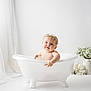 baby, bathtub, clawfoot_tub, headband, white_curtains, flowers, vase, indoor, child, portrait, cute, soft_lighting, smile, infant, clean, decor, white_background, chubby_cheeks, playing, happy
