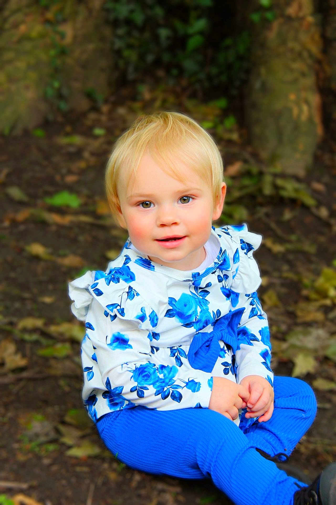 élena a rejoint le concours — aidez-le/la à gagner de superbes lots ! toddler, child, blonde_hair, blue_clothing, floral_pattern, outdoor, forest_floor, tree_trunk, leaves, sitting, cute, portrait, young_child, nature, rosy_cheeks, long_sleeves, pants, casual, innocent, curious