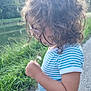 child, curly_hair, glasses, striped_shirt, dandelion, nature, river, grass, path, outdoor, daylight, blue_shorts, greenery, profile_view, casual_clothing, young_kid, summer, peaceful, exploration, curiosity