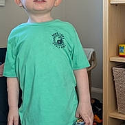 Lathan is registered to the contest to win money with this photo: child, boy, standing, green_shirt, blue_shorts, toy_car, toys, carpet, shelf, basket, indoor, playroom, person, casual_clothing, short_hair, smiling, young, furniture, home, floor