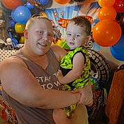 Lathan is registered to the contest to win money with this photo: adult, child, birthday_party, balloons, celebration, indoor, smile, holding, colorful, decorations, person, clothing, shoes, chair, table, toy_car, toddler, happy, family, event
