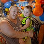 adult, child, birthday_party, balloons, celebration, indoor, smile, holding, colorful, decorations, person, clothing, shoes, chair, table, toy_car, toddler, happy, family, event