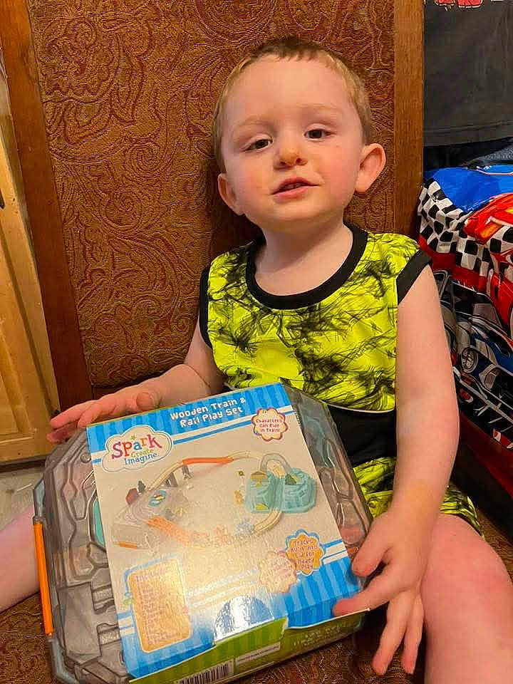 Lathan joined the competition — help win amazing prizes! child, boy, toy, box, wooden_train, rail_play_set, chair, clothing, indoor, furniture, patterned, bedspread, young, sitting, smiling, hand, face, play, yellow, black