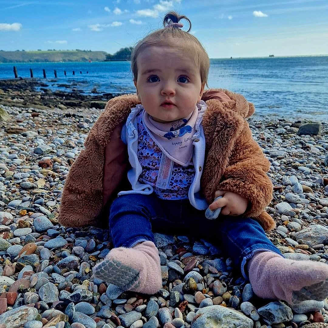 Evie joined the competition — help win amazing prizes! beach, clothing, coast, face, glove, head, nature, outdoors, pants, pebble, person, photography, portrait, rock, sea, shoreline, sitting, sky, water, waterfront