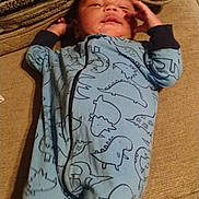 Ryder is registered to the contest to win money with this photo: baby, infant, blue_onesie, dinosaur_pattern, couch, cushion, indoor, child, sleepy, relaxed, hand_near_face, textile, fabric, portrait, person, home, furniture, soft_lighting, infant_clothing, resting