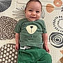 baby, child, smiling, green_clothing, tshirt, pants, lying_down, patterned_mat, indoor, cute, happy, face, person, infant, toddler, short_hair, hand, footwear, text_on_clothing, playmat