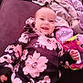 baby, smiling, floral_clothing, hoodie, plush_toy, blanket, couch, indoor, child, happy, face, person, infant, cute, soft_toy, portrait, lying_down, cozy, colorful, playful