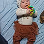 Leenna is registered to the contest to win money with this photo: baby, infant, toy, blue_sheet, white_hat, brown_skirt, leggings, socks, chewing, lying_down, indoor, child, playing, cute, soft_lighting, patterned_clothing, toddler, smiling, happy, colorful_toy