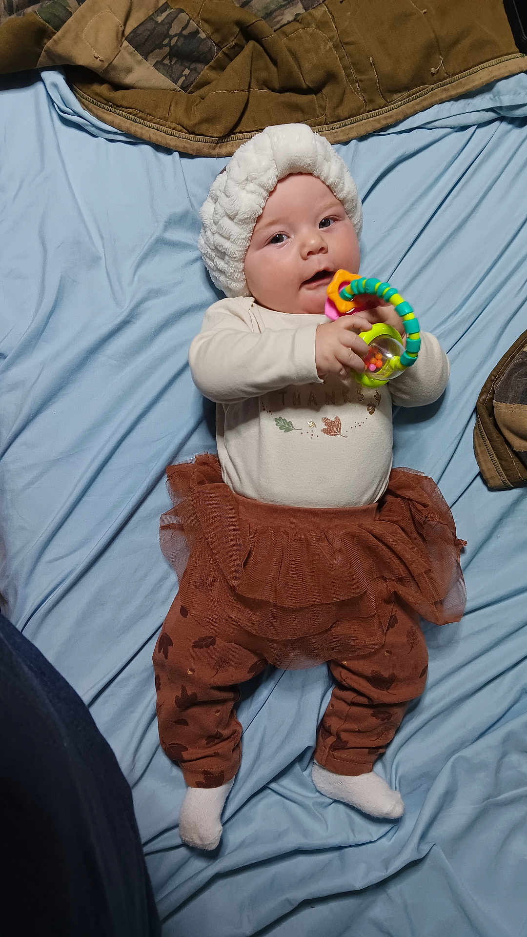 Leenna is registered to the contest to win money with this photo: baby, infant, toy, blue_sheet, white_hat, brown_skirt, leggings, socks, chewing, lying_down, indoor, child, playing, cute, soft_lighting, patterned_clothing, toddler, smiling, happy, colorful_toy