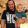 man, baby, smile, indoor, store, clothing, green_shirt, socks, holding, happy, person, face, long_hair, beard, goatee, sign, lighting, background, retail, family
