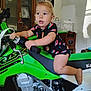 toddler, motorcycle, indoor, child, green, toy, bike, vehicle, face, hands, legs, black_clothing, patterned_clothing, metal, engine, furniture, cabinet, room, curious, grip