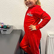 Amelia is registered to the contest to win money with this photo: barefoot, casual, chair, child, face, furniture, girl, hair, hand_on_hip, happy, indoor, jumpsuit, person, pose, radiator, red_clothing, room, smiling, table, wall